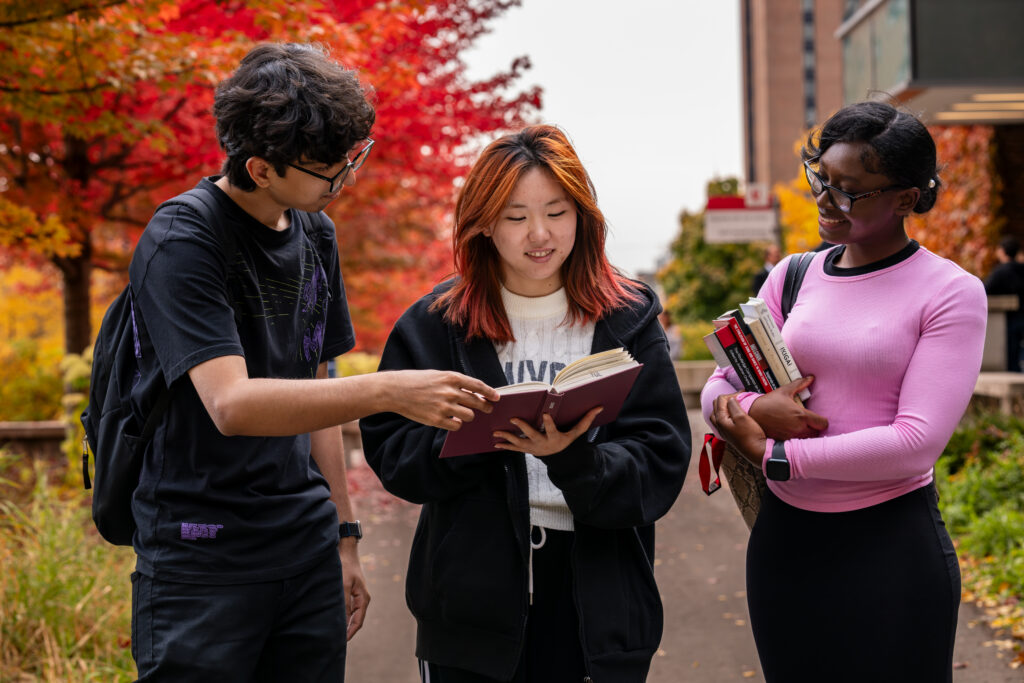 Three university students stand on a campus pathway lined with vibrant red and orange autumn trees; one student points to a page in a notebook held by another, while the third smiles and holds a stack of books.