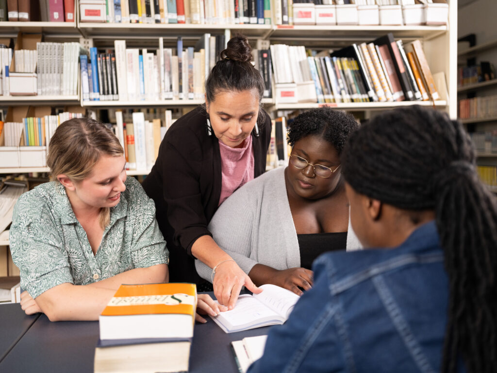 A group of three students sit around a table in a library as an instructor stands beside them, pointing to a page in an open book. The students are engaged in the discussion, surrounded by books and shelves filled with academic texts.