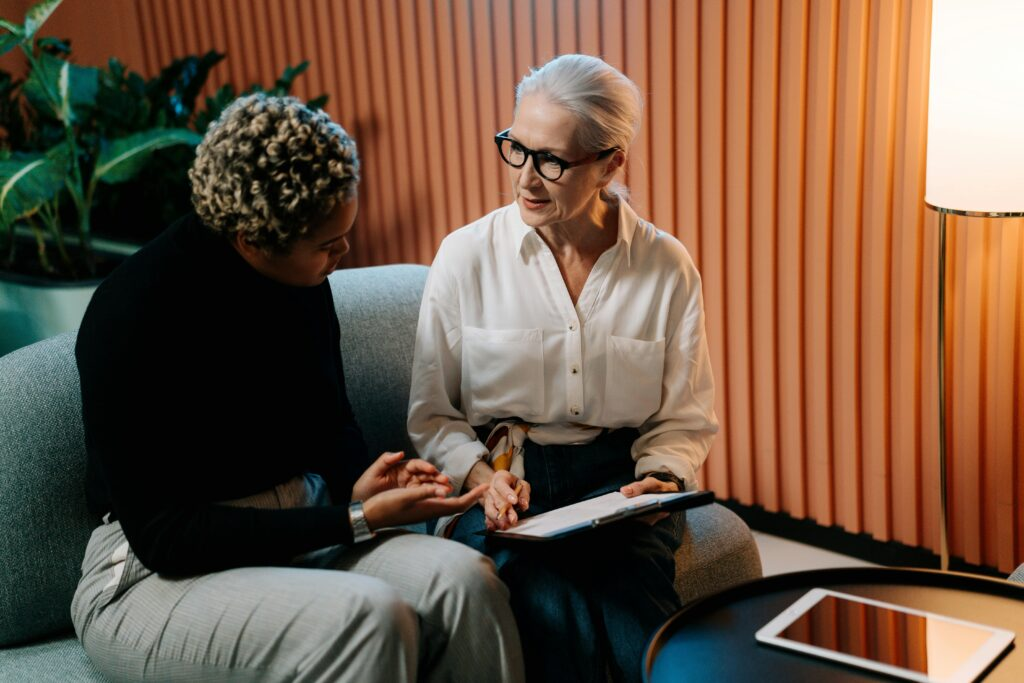 Two women sit on a blue couch engaged in a conversation. One, with short curly hair, wears a black top and gray pants; the other, with white hair and glasses, wears a white blouse and holds a clipboard. A tablet rests on a nearby table, and the setting includes warm lighting and a contemporary interior with wooden paneling and potted plants.