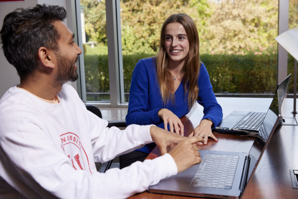 Two graduate students sit at a table with laptops, engaged in conversation, with natural light streaming through large windows and greenery visible outside.