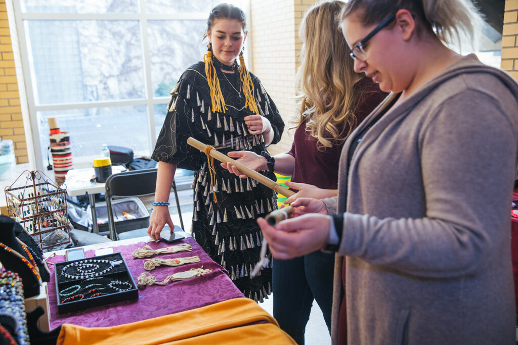 Three women stand at a display table featuring Indigenous beadwork and cultural items; one, dressed in traditional clothing shares information while the others examine handcrafted objects.