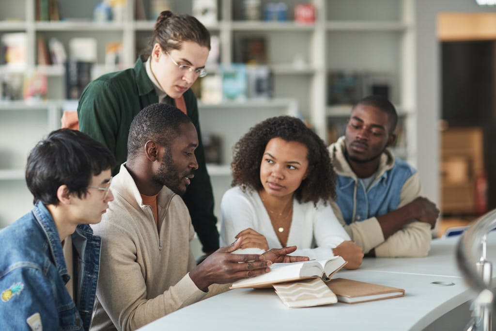 A group of five people sitting around a table in a library or study space, engaged in a discussion. One person is speaking while gesturing towards an open book, and the others are listening. Books and notebooks are spread out on the table. Shelves filled with books are visible in the background. 