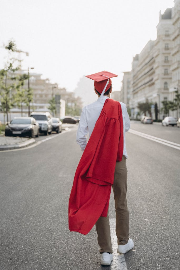 A graduate wearing a red cap and carrying a matching red graduation gown walks down the middle of a city street. The individual is seen from behind, dressed in casual pants and a white shirt, with tall buildings and parked cars lining both sides of the road.