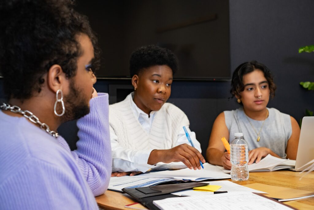 Three graduate students sit around a table in discussion, with open books, notebooks, and highlighters in front of them; one wears a lavender sweater, another person in a white sweater gestures with a pen, and the third person sits beside a laptop and water bottle.