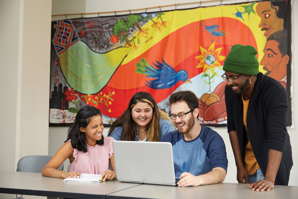 Three students are sitting at a table and 1 student is standing. They are all looking at a laptop and smiling. There is a colourful wall hanging in the background.