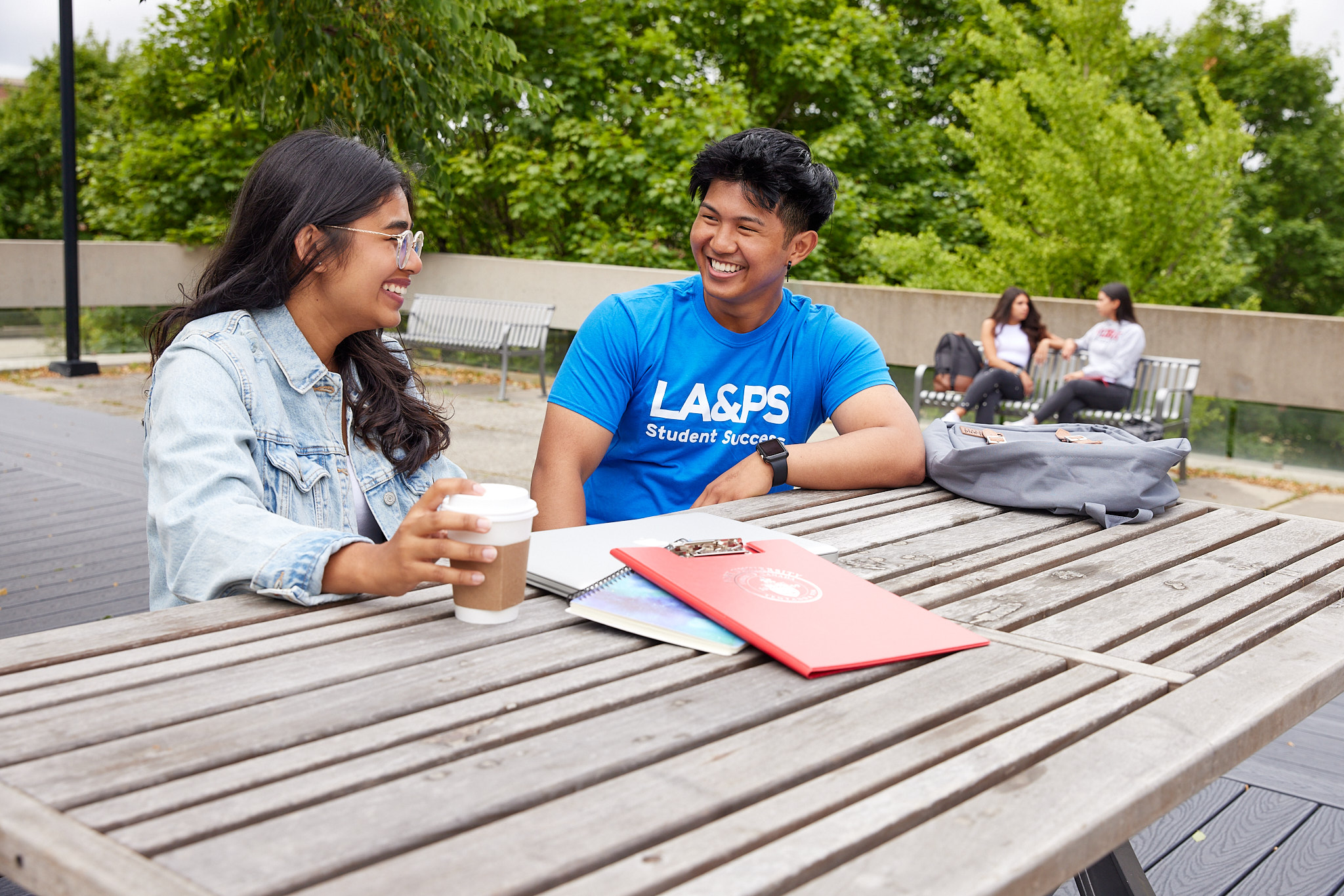 Two university students are sitting at a picnic table outdoors and there is a knapsack and red clipboard and coffee cup on the table. One student is wearing a blue LA&PS tshirt and one student is wearing a light blue jean jacket.