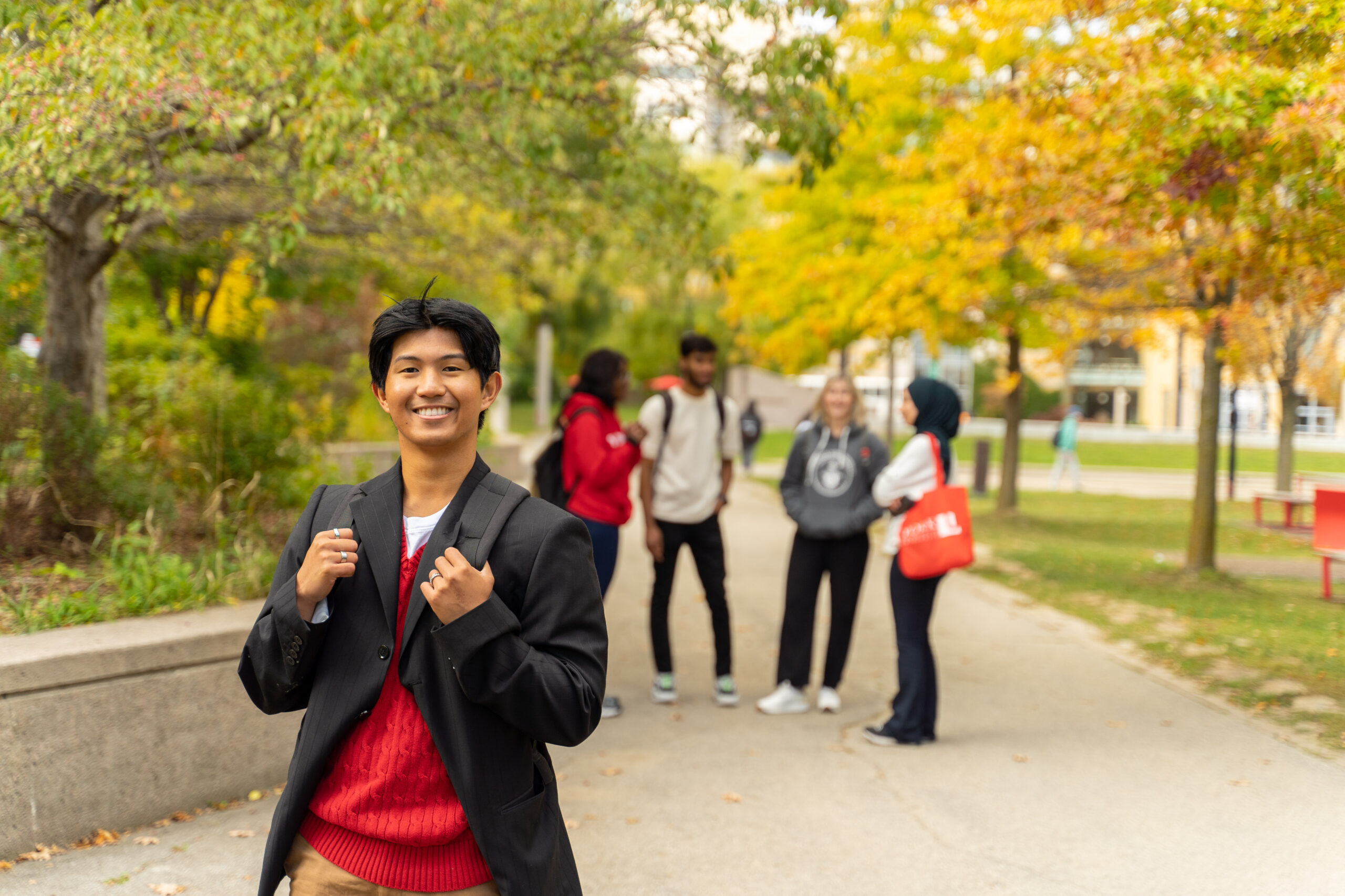 A student in the foreground facing the camera smiling and wearing a red sweater and black jacket with a knapsack. There are 4 students standing in the background that is blurred.
