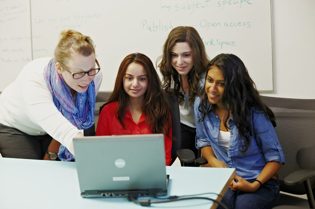 A group of 3 students and 1 teacher in a classroom setting. They are all looking at a laptop on a desk and smiling. There is a whiteboard in the background.