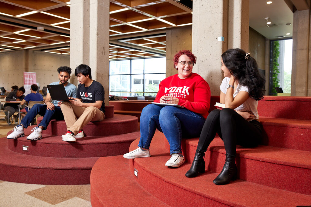 Four YorkU students sitting indoors on carpet-covered steps. Two students on the right side are facing each other and smiling. Two students on the left side are looking at an open laptop.