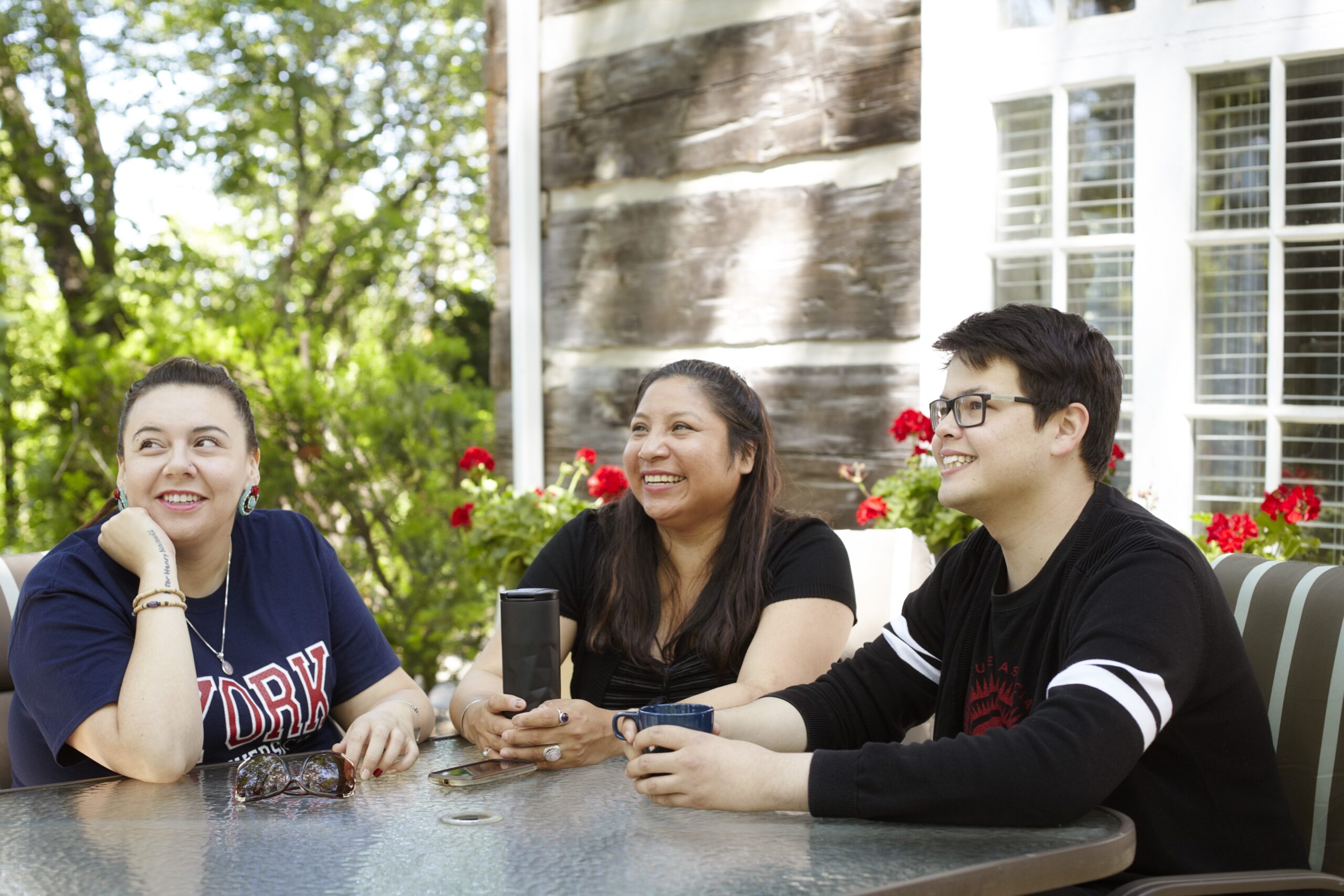Three people in an outdoor setting are seated at a table with trees, red flowers and a wood panelled house in the background. One person is holding a blue tea cup, one person is holding a black travel mug and one person has a hand on a cheek with sunglasses on the table in front.