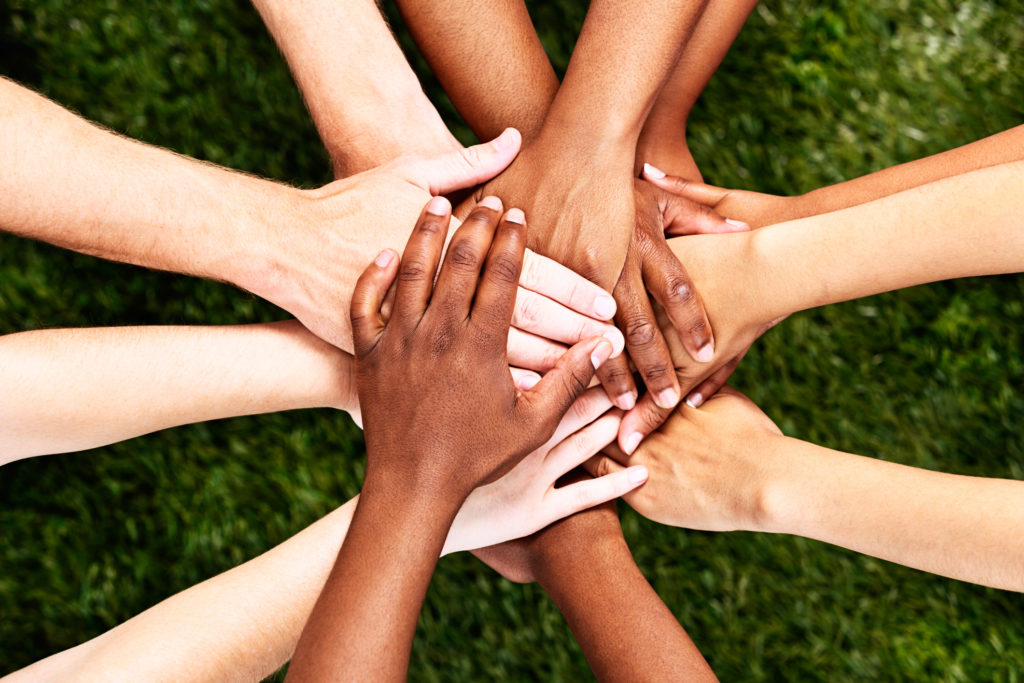A pile of multiracial hands are stacked in support or unity, against a background of grass. All for one and one for all!
