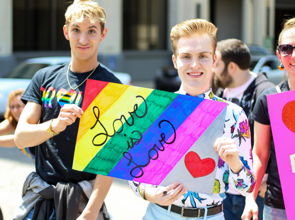 photo of two people holding a homemade Love is Love sign with a rainbow-coloured background