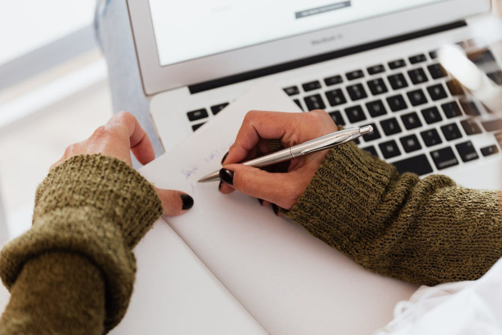 image of hands writing with a pen and holding paper over a laptop