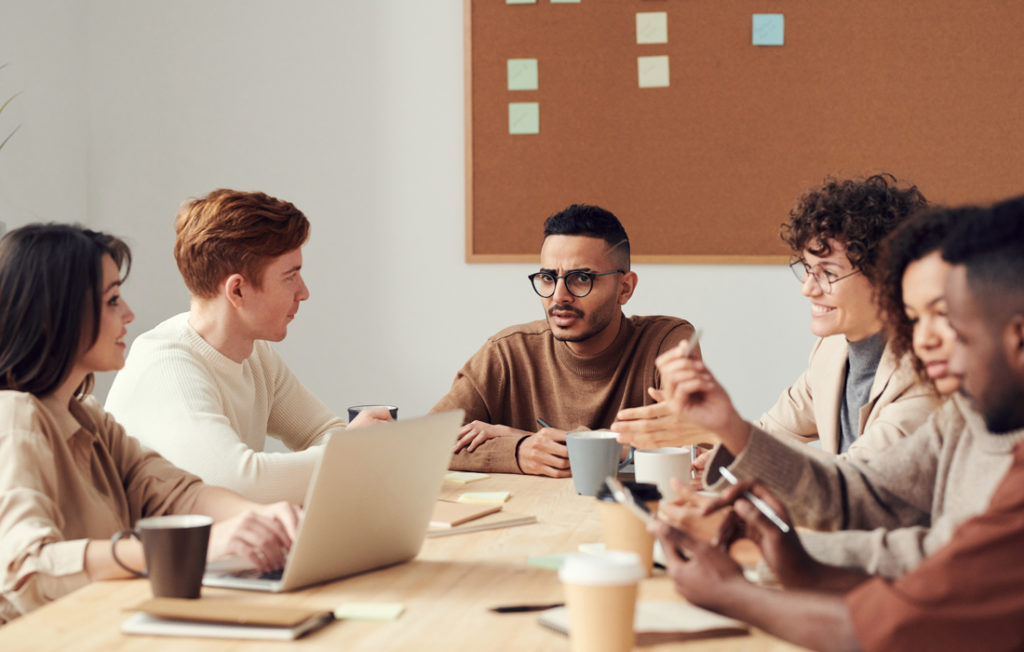 six people sitting at a table conversing, one has a laptop and one is using a phone