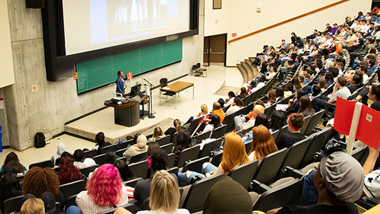 students inside large lecture hall listen to a professor deliver a lecture.