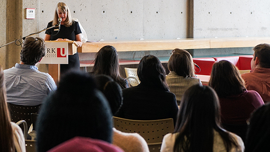 picture showing back of individuals heads who are sitting and attending a lecture with a woman speaker at the podium