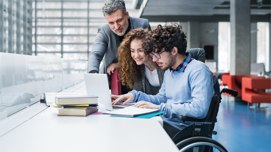 person in wheelchair using computer and two people beside him