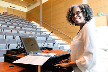 a Black woman professor turned to her side smiling, while the lecture hall seats are empty, she has a laptop and a folder open in front of her