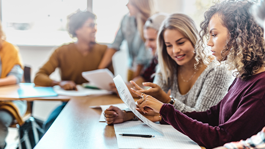 Four students sit and one stands around a table going over an assignment together.