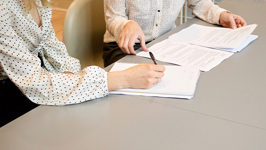 A person sits at a table explaining some forms to another person who is taking notes.