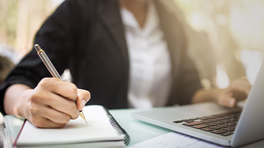 executive female sitting at a desk writing in a notebook in front of laptop
