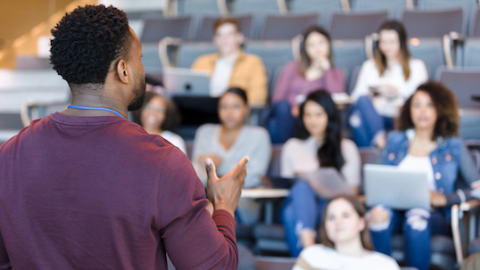 A male college professor gestures while giving a lecture to a group of college students.