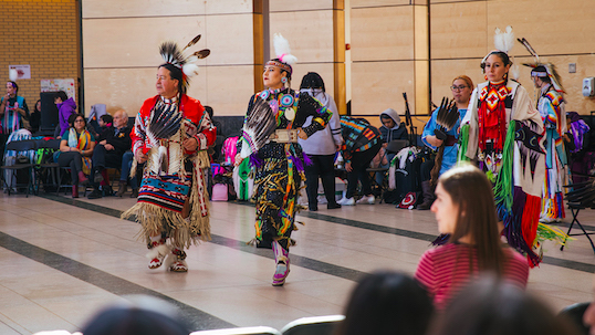 Man and two women wearing Indigenous attire while performing in front of Vari Hall crowd at Keele Campus
