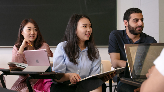 Three students are seated in a classroom and participating in a lecture using laptops. 