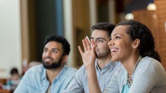 Three students are seated in a classroom and participating in a lecture by raising their hands and asking questions.
