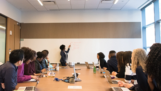 A female worker gives a conference presentation in a boardroom while her colleagues watch. 