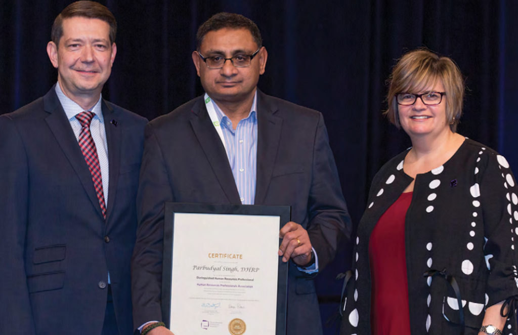 A man holds up an award while two other people, a man to his left and a woman to his right, both smiling, surround him for a photo opportunity. 