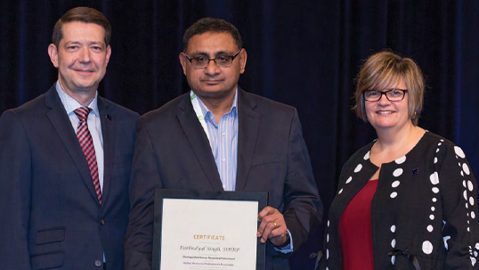 A man holds up an award while two other people, a man to his left and a woman to his right, both smiling, surround him for a photo opportunity. 