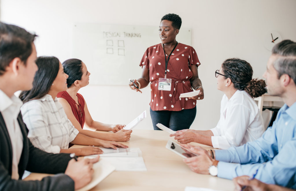 A woman is giving a presentation to her co-workers while holding notes in a conference room. He coworkers are gather around a table taking notes as they listen to her.