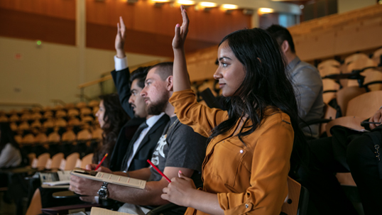 Three students are seated in a classroom and participating in a lecture by raising their hands and asking questions.