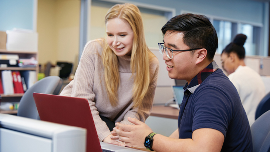 Male staff member helps female student with work on her computer in a university classroom. 