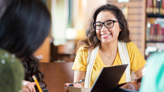 A female university student smiles at a fellow student as they work on an assignment in a university library. 