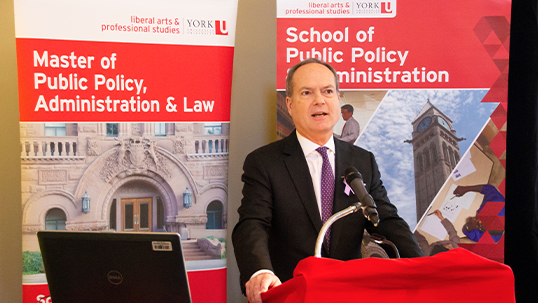 Man in formal attire stands at podium while giving presentation and standing in front of York University public policy program branded posters