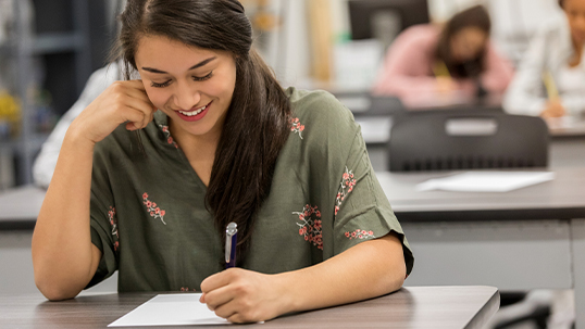 Female student writes on paper while sitting down at desk