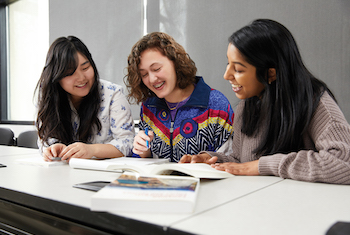 Three students smiling and looking down at a book on a table