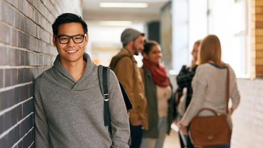 A student wearing a grey sweater and glasses leaning against a wall