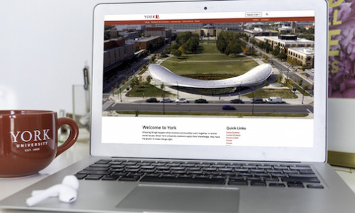 A closeup shot of a laptop on the Welcome to York homepage. A red York coffee mug is beside it and a pair of wireless headphones rest on the laptop