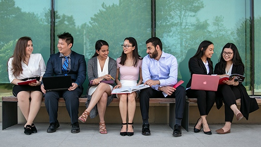 A group of university students dressed professionally are seated outside and are discussing their next lecture before entering class