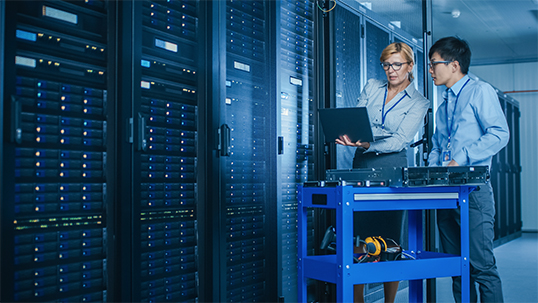 woman standing with male student in server room showing something on computer