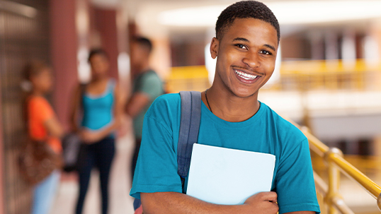 male student in blue shirt holding books at chest, people in background blurred