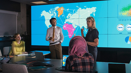 two people standing, two people sitting, discussing in front of map of the world on tv screens