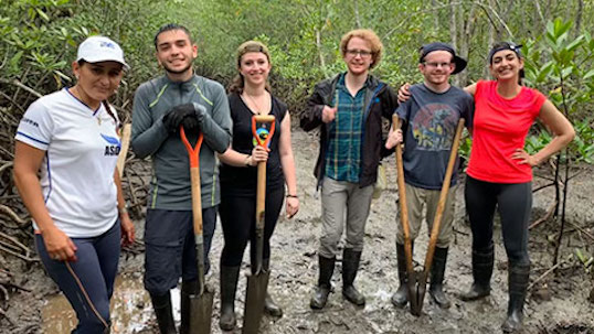 Six students pose for happy photo while holding shovels in muddy outdoor setting