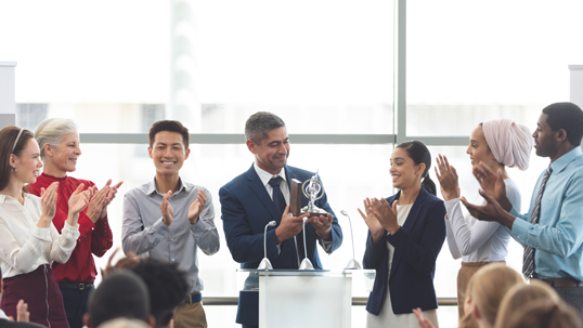 Multicultural group of men and women applaud man in suit and tie accepting an award