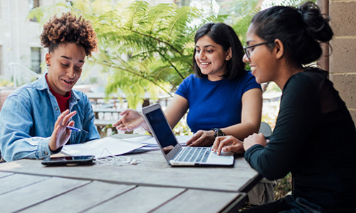 three diverse students sit on a lawn table and smile while they discuss work with a laptop and notes open