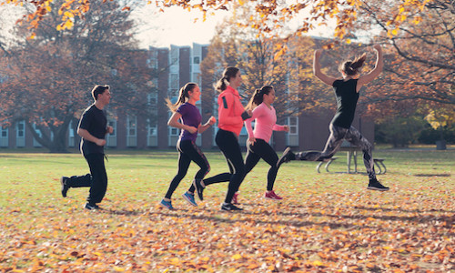 five members of a club in athletic attire running in yorku lawn with one woman in front taking a big leap