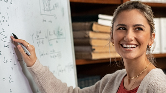 young woman smiles while writing algebraic equations on whiteboard